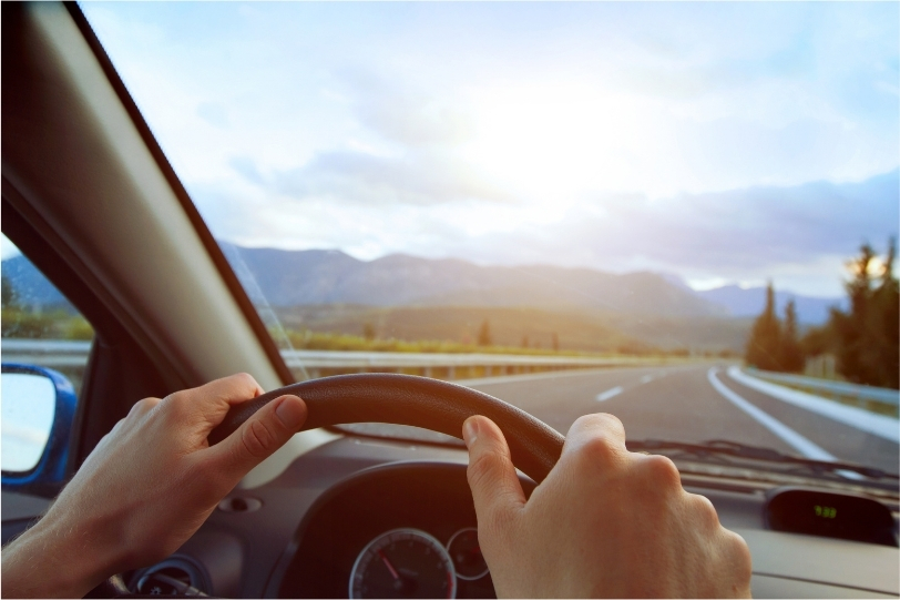 Deux mains de conducteur sur un volant devant une autoroute ciel bleu et soleil, montagnes au loin et paysages de foret